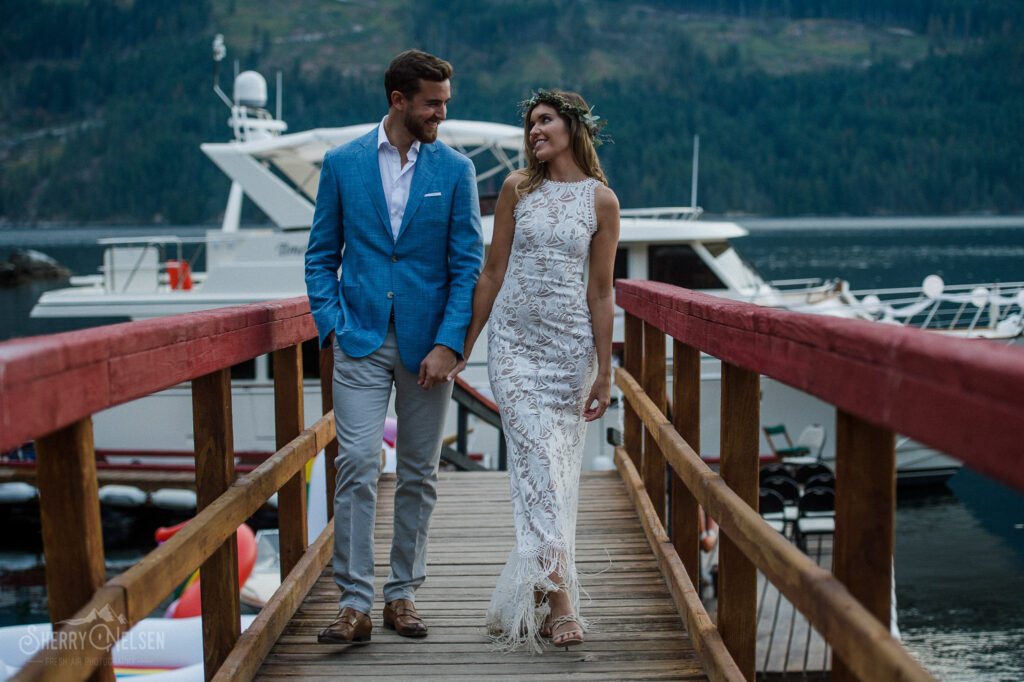 a young bride wearing a close fitting lace gown and a flower crown walks smiling down the pier in sechelt bc with her new husband who wears a blue sport jacket and grey pants