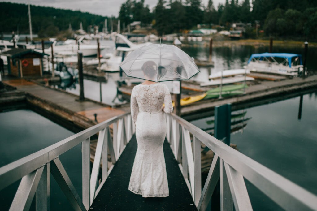 a sunshine coast bc bride walks down a ramp to a marina on a rainy day with a clear umbrella covering her from the rain