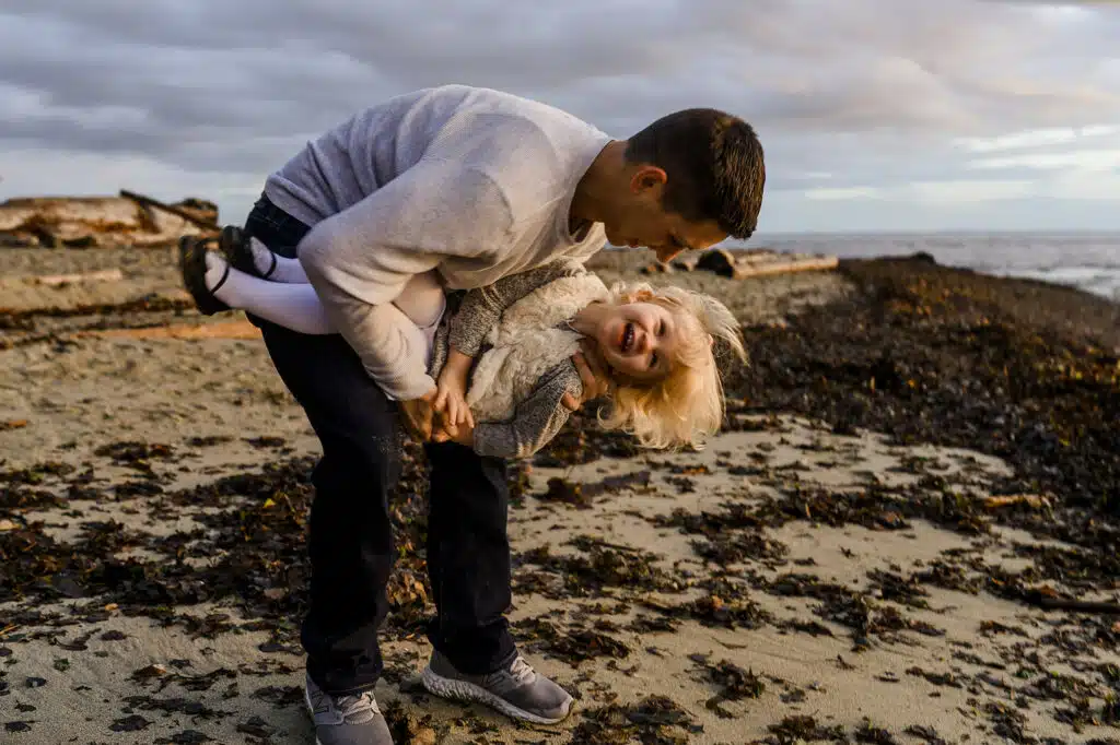 a young father plays with his blonde toddler daughter on the beach at davis bay in sechelt bc