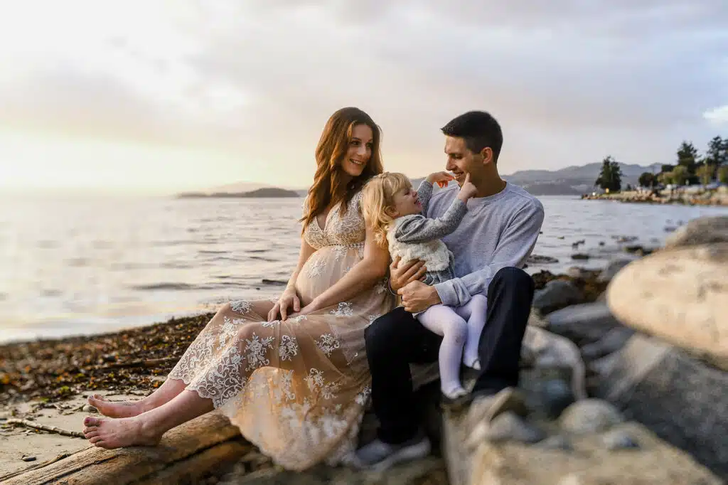 a pregnant woman and her husband sit on the beach at davis bay on the sunshine coast of bc with their young daughter during a family photo session with fresh air photography